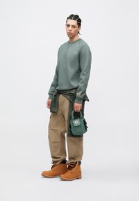 Žalias džemperis, chaki kelnės ir rudi batai. Aprangoje yra žalia utilitarinė rankinė ir tekstūruotos medžiagos detalė ant džemperio.