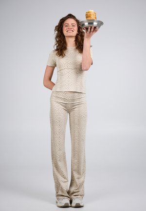 Kvinde iført beige leopardprint-outfit, der holder en metalplade med en høj stak pandekager og smiler mod en ensfarvet baggrund.