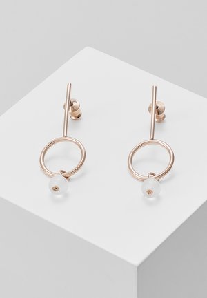 Boucles d'oreilles en métal doré avec une barre verticale, un cercle ouvert et une petite perle blanche suspendue à l'intérieur de chaque cercle, présentées sur une surface blanche.