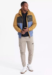 Färgblockerad jacka i tan, blå och svart; har huva, dragkedja och logotyp. Matchas med khaki cargobyxor och vita sneakers.