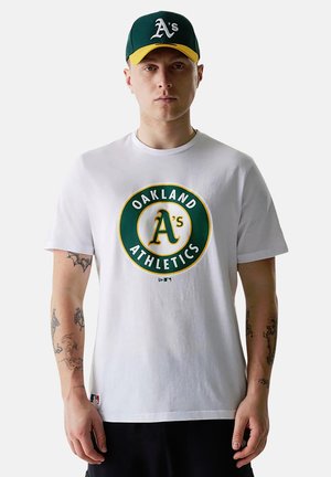 Hombre con camiseta blanca de Oakland Athletics y gorra de béisbol verde y amarilla, de pie con expresión neutral frente a un fondo liso.