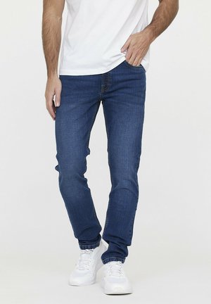 Blaue Denim-Jeans mit tailliertem Schnitt, Fünf-Taschen-Stil und dezenten Stichdetails, kombiniert mit weißen Sportschuhen und einem weißen T-Shirt.