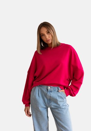 Kobieta ubrana w jasnoczerwoną, oversize'ową sweterkę i jasnoniebieskie jeansy z wysokim stanem, stojąca z jedną ręką w kieszeni na białym tle.