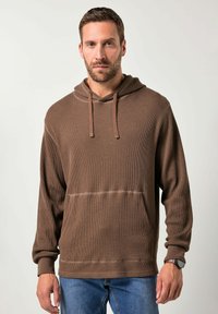 Bruine hoodie gemaakt van gestructureerde stof, met een voorvak en geribbelde manchetten. Gecombineerd met blauwe jeans.