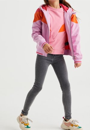 Vaate takki vaaleanpunaisena ja oranssina vetoketjulla, jossa huppu, yhdistetty vaaleanpunaiseen collegepaitaan, harmaisiin tiukkoihin farkkuihin ja monivärisiin tennareihin.