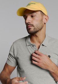 Grijze polo met drie knopen en een klein zwart logo op de borst. Gele pet met groene borduring. Close-up weergave.