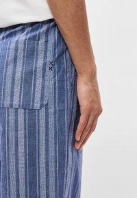 Ριγέ μπλε παντελόνι από λινό ύφασμα. Περιέχει μία πλαϊνή τσέπη με εναλλασσόμενο μπλε ραφές. Οι κατακόρυφες ρίγες διαφέρουν σε πλάτος.