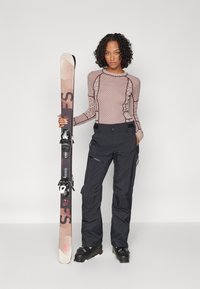 Ski outfit featuring a patterned long-sleeve top in mauve and black pants with zippers. Ski with a pink and black design held in left hand.