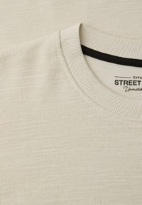 Col rond beige texturé avec une doublure intérieure noire et un texte partiellement visible « STREET » sur l'étiquette à l'intérieur du col.