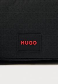 Borsa in tessuto nero con motivo "Hugo" in rilievo e logo rosso prominente su etichetta in pelle nera. Texture liscia con design elegante.