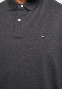 Dark gray polo shirt made of cotton with a collar, featuring two white buttons and a small red, white, and blue logo on the chest.