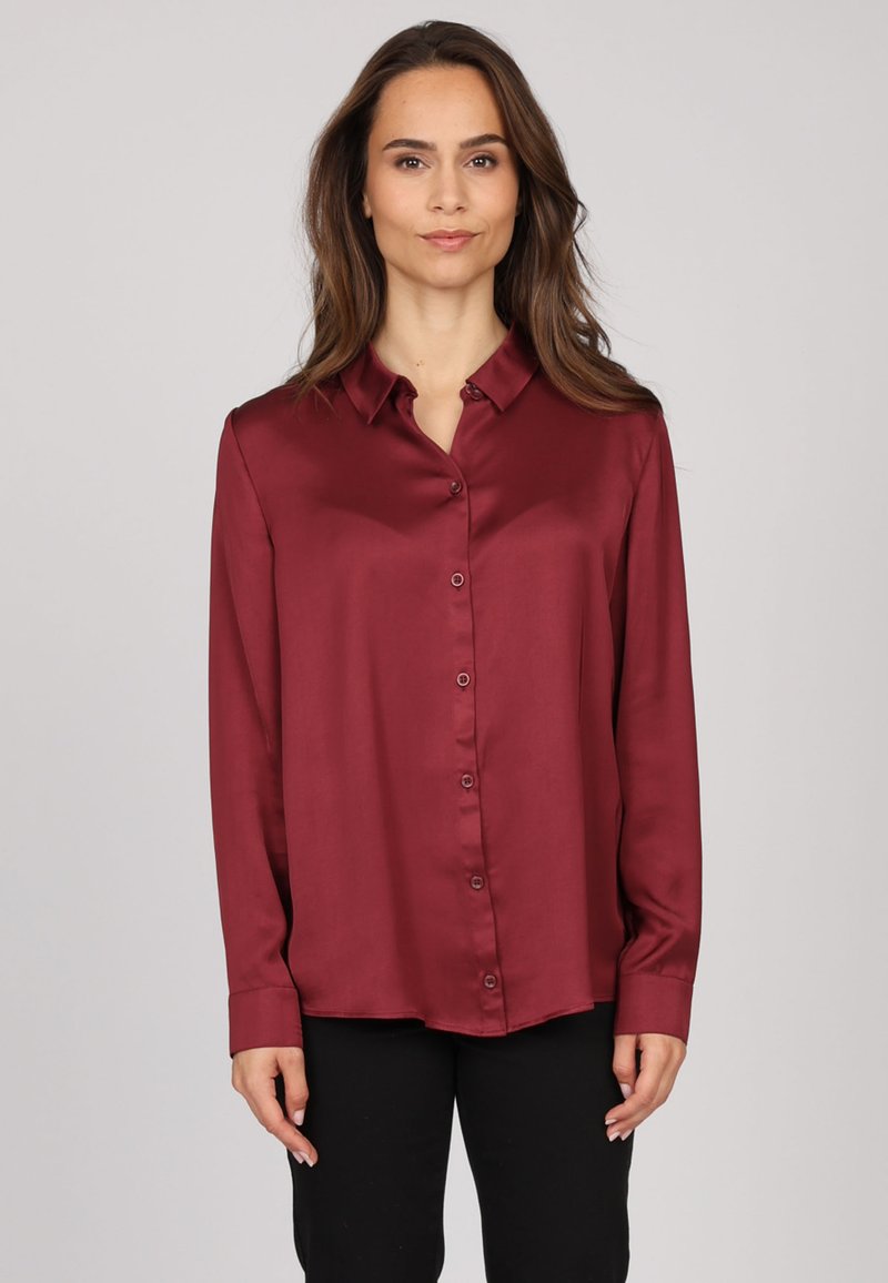 Chemise en soie bordeaux à boutons, avec un col, des manches longues et une texture lisse. Présente des boutons sur le devant et des poignets ourlés.