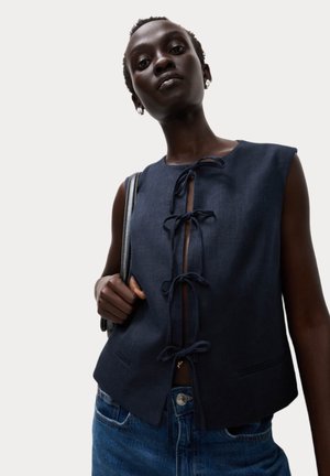 Gilet sans manches bleu marine avec un lien à l'avant, fabriqué à partir d'un tissu texturé. Présente un léger crop et des poches, porté avec du denim.