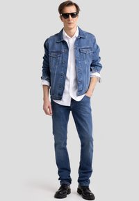 Férfi napszemüvegben, kék farmerdzsekiben, fehér ingben, kék farmernadrágban és fekete csizmában, egyik kezét a zsebében tartva áll, szürke háttér előtt.