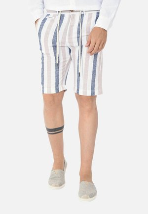 Uomo che indossa pantaloncini al ginocchio con righe verticali beige, bianche e blu, scarpe slip-on grigie e un tatuaggio nero a doppia fascia sulla gamba.
