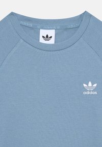 Lys blå ribbestrikket t-skjorte med hvit Adidas-logo. Har raglan-ermer og en myk tekstur.