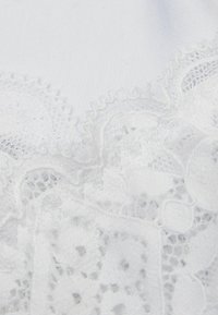 Tissu en dentelle blanche avec des motifs floraux et des détails complexes, contrasté contre un fond blanc lisse, mettant en valeur la texture et le design.