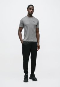 Szara koszulka z okrągłym dekoltem z małym czarnym logo, zestawiona z czarnymi zwężanymi dresami i czarnymi trampkami, wszystko na jednolitym białym tle.