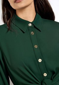 Grünes Hemd mit einem spitzen Kragen, ausgestattet mit goldfarbenen Knöpfen und einem geknoteten Detail in der Taille. Glatte Stoffstruktur.