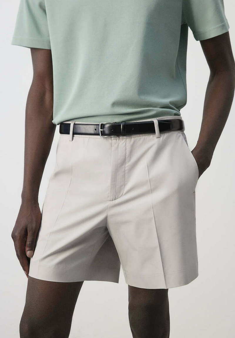 Person wearing light gray tailored shorts with front pleats, black belt, and tucked-in light green short sleeve shirt, standing with one hand in pocket.