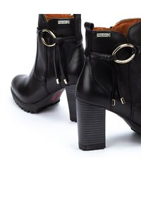 Botas de tobillo de cuero negro con punta cuadrada, tacón cilíndrico, banda decorativa con un acento circular y suela texturizada.