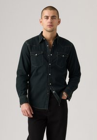 Dark green denim shirt with pointed collar, two chest pockets, and contrasting yellow stitching. Styled with black pants, worn unbuttoned at the collar.