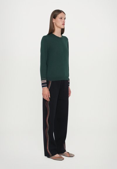 Pulover verde tricotat cu mâneci lungi și manșete împodobite, purtat cu pantaloni largi bleumarin cu o dunga colorată pe lateral și pantofi fără toc.