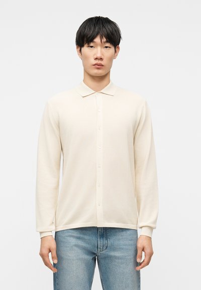 Jeune homme aux cheveux noirs portant un polo à manches longues crème boutonné et un jean bleu clair, debout devant un fond uni.