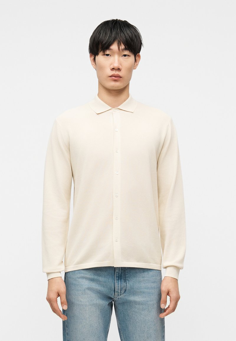 Jeune homme aux cheveux noirs portant un polo à manches longues crème boutonné et un jean bleu clair, debout devant un fond uni.