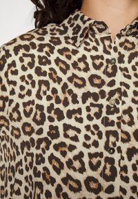 Chemise à boutons en imprimé léopard avec un fond beige clair, des taches brunes foncées et un col classique. Présente un léger reflet et une texture lisse.