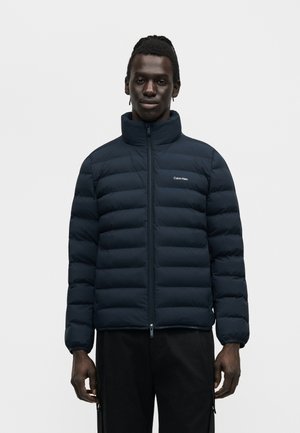 Mann trägt eine schwarze Steppjacke mit Stehkragen und Calvin Klein-Logo auf der Brust, steht vor einem einfarbigen Hintergrund.