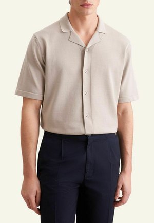 Uomo che indossa una camicia beige a maniche corte con bottoni e colletto e pantaloni blu scuro su uno sfondo chiaro uniforme.