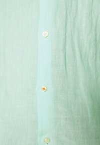 Jasnozielona tkanina lniana z białymi guzikami, jeden guzik przyszyty pomarańczową nitką.