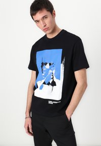 Camiseta negra de algodón con un estampado gráfico azul y blanco de una cara y el texto "KARL LAGERFELD JEANS" en la parte inferior.