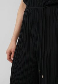 Schwarzer plissierter Jumpsuit mit eng anliegendem Oberteil, dünnem Gürtel in der Taille und goldenen Akzenten an den Gürtelenden, der eine glatte Textur und ein strukturiertes Design präsentiert.