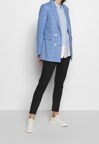 Blazer a quadri blu chiaro con bottoni dorati, camicia a righe bianche, pantaloni neri slim-fit e sneakers crema, su uno sfondo semplice.