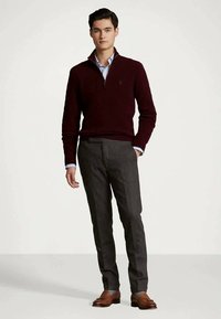 Młody mężczyzna stoi w bordowym swetrze z krótkim zamkiem, jasnoniebieskiej koszuli, szarych spodniach i brązowych mokasynach na jednolitym tle.