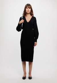 Rochie tricotată neagră, cu decolteu în V, mâneci lungi și voluminoase, și siluetă ajustată, care ajunge până la jumătatea gambei. Accesoriile includ o geantă de mână neagră și balerini.