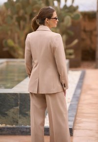 Woman with brown hair in a ponytail wearing sunglasses and a beige suit stands near a pool with cactus plants in the background.