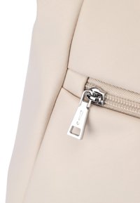 Zaino in tessuto beige con una trama liscia. Presenta un cursore in metallo argentato con un simbolo di avviso triangolare e un'apertura rettangolare.