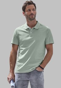 Polo verde chiaro in cotone, con colletto classico e patta con tre bottoni, indossato con jeans grigi. Dettaglio del materiale testurizzato visibile.