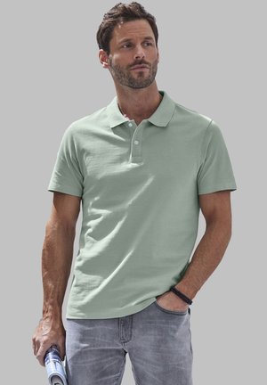 Polo verde chiaro in cotone, con colletto classico e patta con tre bottoni, indossato con jeans grigi. Dettaglio del materiale testurizzato visibile.