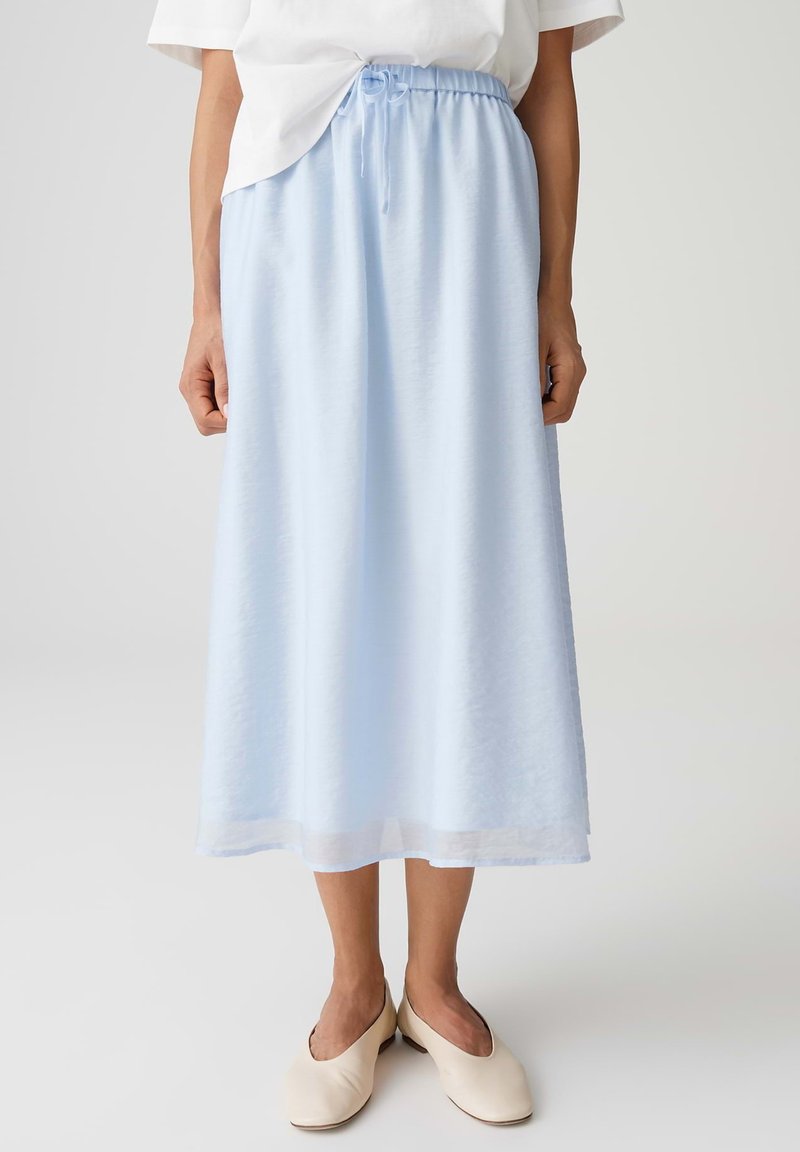 Hellblauer Midirock mit elastischem Bund und Kordelzug, kombiniert mit cremeweißen flachen Schuhen und einem in den Rock gesteckten weißen Oberteil.