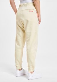 Cremfarbene Sweatpants mit einem Kordelzugbund, elastischen Bündchen und einer hinteren Reißverschlusstasche mit einem leuchtend pinken Logo.