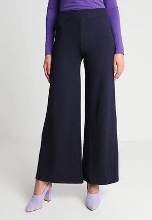 Vrouw die zwarte broek met wijde pijpen draagt, paarse top met lange mouwen en lavendelkleurige schoenen met blokhak, staand tegen een neutrale achtergrond.