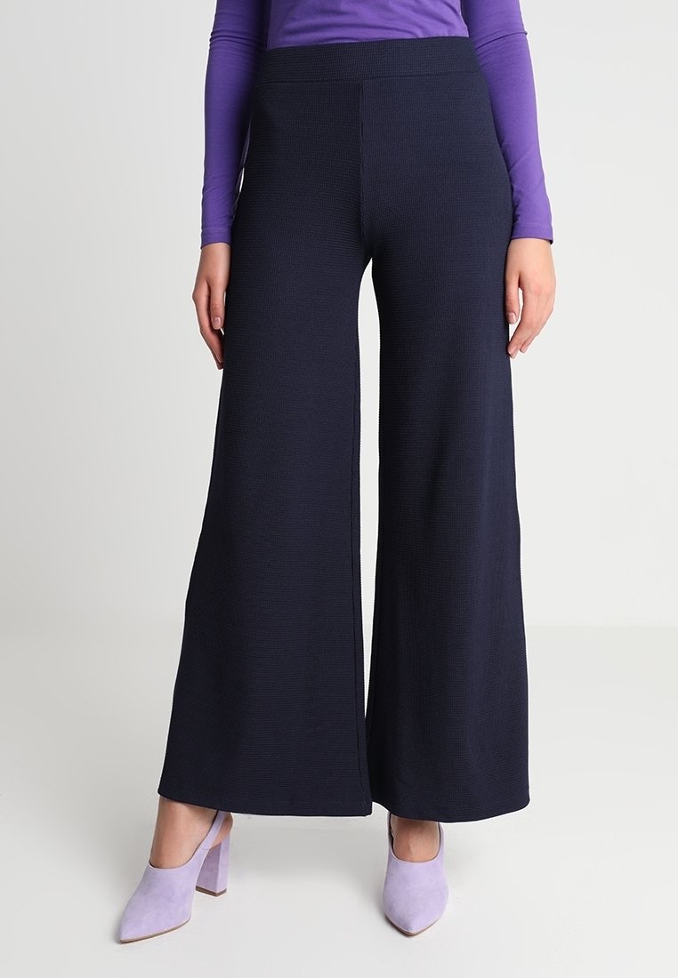 Femme portant un pantalon noir à jambes larges, un haut violet à manches longues et des chaussures à talons blocs lavande, debout devant un fond neutre.