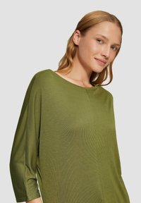 Groene, lange mouwen top van textuurstof, met een brede halslijn en een iets relaxte pasvorm. Zijsplitten voegen een moderne toets toe.