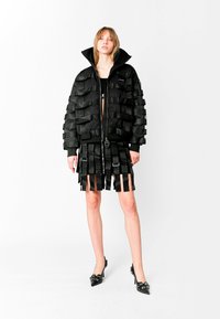 Chaqueta acolchada negra con detalles de cintas en capas con acabado satinado, cuello alto, cierre de cremallera; combinada con una falda corta plisada de cintas. Tacones altos negros.