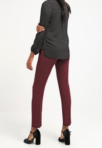Blusa nera a maniche lunghe con spacchi laterali abbinata a pantaloni aderenti color bordeaux e scarpe nere con tacco a blocco, vista da dietro.