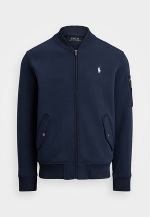 Veste bomber bleu marine en tissu doux, dotée d'une fermeture éclair à l'avant, de deux poches latérales, d'une poche sur la manche avec fermeture éclair et d'un logo blanc sur la poitrine.